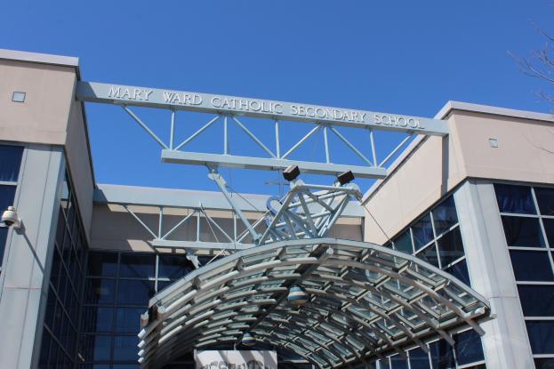 Mary Ward Catholic Secondary School building with glass walls, a name board, an arch with lights, a banner, a tree on the right, and a clear blue sky.