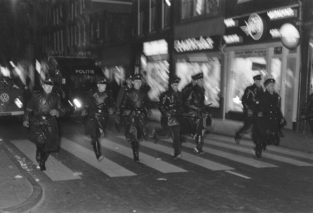 Polizisten in Uniform und mit Mützen bei Nacht über eine Straße gehend, einige mit Waffen, mit Fahrzeugen und Gebäuden im Hintergrund in Schwarz-Weiß.