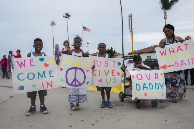 Eine Gruppe von Kindern mit Schildern vor einem Gebäude, mit einer Frau, die einen Kinderwagen mit einem Baby schiebt, auf einer Straße umgeben von Bäumen, Pfählen, einer Flagge, einem Haus, Pflanzen und einem bewölkten Himmel; auf einem Schild steht: "Wir kommen in Frieden, gebt uns unseren Daddy."