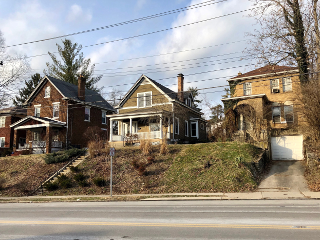 Reihe von Häusern mit Fenstern, Türen, Säulen, Geländern, Stufen, Pflanzen und einem Pfahl mit einer Tafel, vor einem bewölkten Himmel.