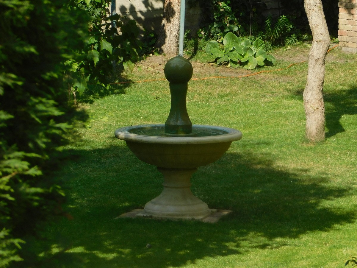 Ein Garten mit einem zentralen Brunnen, umgeben von grünem Gras und Bäumen und einer Wand mit Pflanzen im Hintergrund.