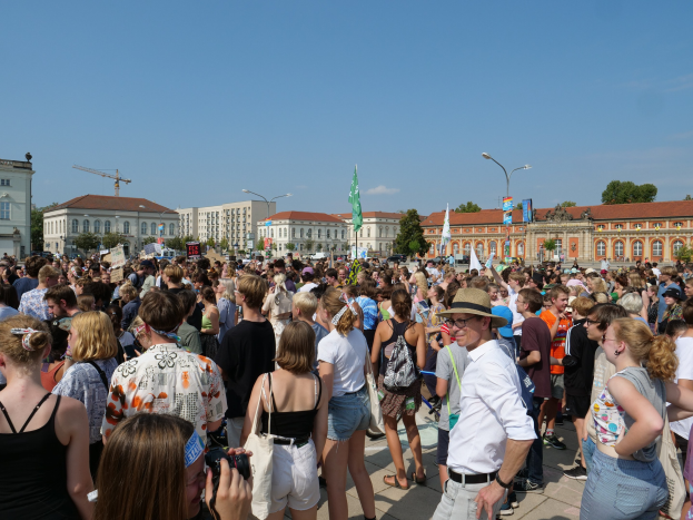 Große Menschenmenge vor einem Gebäude mit Fahnen, Bäumen und einem bewölkten Himmel, einige halten Kameras während einer Demonstration.