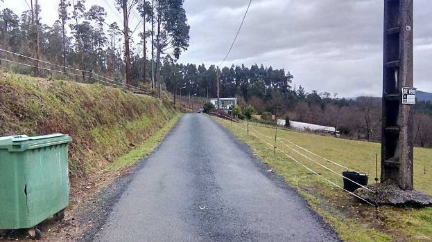 Grüner Mülltonnen am Rande einer Straße mit Gras, Pflanzen, Bäumen, Masten, Drähten und einem Zaun, unter einem bewölkten Himmel, mit einer Masttafel auf der rechten Seite und verstreuten Gegenständen auf der linken Seite.