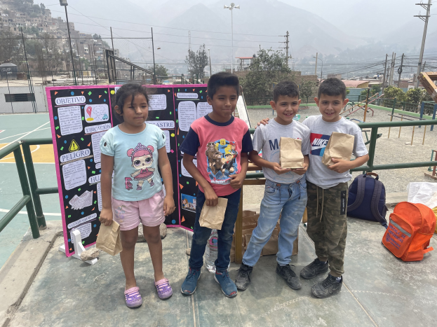 Gruppe von Kindern, die Papiertüten auf einem Basketballfeld halten, mit Tüten und Gegenständen auf dem Boden, einer Tafel mit Text links und Zäunen, Pfählen und Gebäuden im Hintergrund.