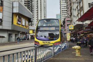 Ein gelber Doppeldeckerbus fährt eine Straße mit hohen Gebäuden entlang, mit Menschen auf dem rechten Gehweg, einem Feuerhydranten und einem Geländer, unter einem klaren blauen Himmel.