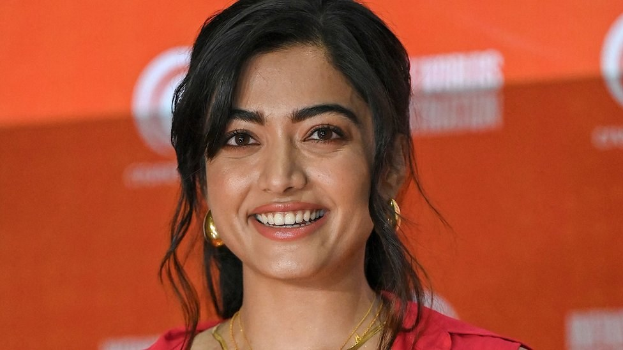 Eine Frau in einem roten Kleid mit einem eleganten Dutt und dezentem Make-up steht lächelnd da, trägt schwarze High Heels und hat ein hellgelbes Banner im Hintergrund mit der Aufschrift 'Sonam Kapoors Schönste Looks vom Cannes Film Festival'.