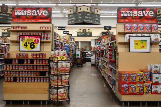 Ein Kiosk-Innenraum mit Regalen und Boden voller verschiedener Lebensmittel, beleuchtet von Deckenlampen und mit Informationsschildern mit Text und Zahlen im Hintergrund.