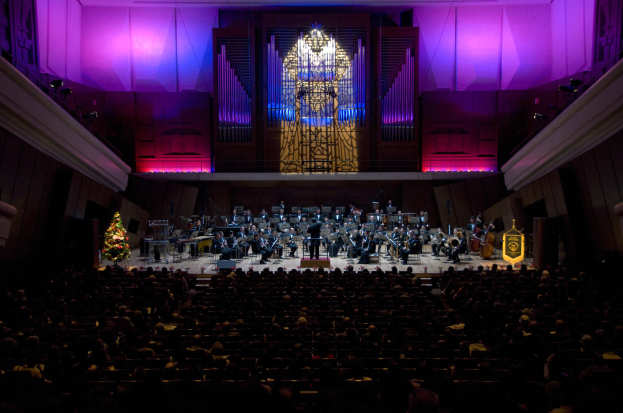 Großer Saal mit Menschen, die ein Konzert auf der Bühne verfolgen, mit einem Weihnachtsbaum und geschmückten Wänden.