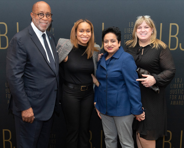 Eine Gruppe von vier Personen auf einem roten Teppich, die fröhlich posieren und fotografiert werden, mit einem 'Liberty Justice for All Awards'-Schild im Hintergrund.