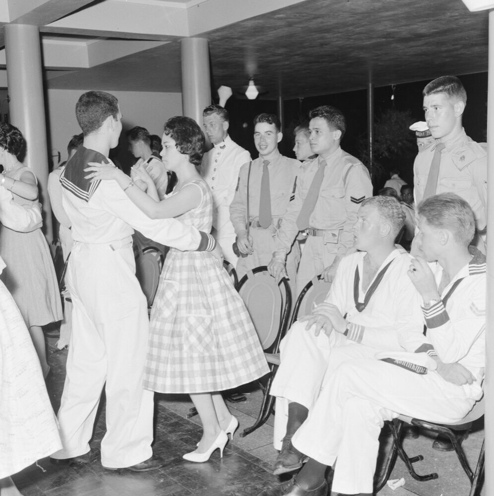 Schwarz-weiß-Bild einer Gruppe von Menschen in Militäruniformen, die auf einer Tanzfläche tanzen und sich vergnügen, mit Säulen und Lichtern im Hintergrund.