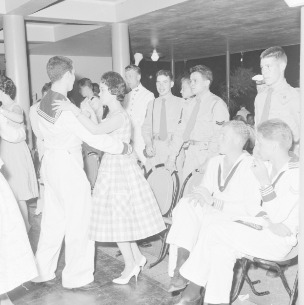 Schwarz-weiß-Bild einer Gruppe von Menschen in Militäruniformen, die auf einer Tanzfläche tanzen und sich vergnügen, mit Säulen und Lichtern im Hintergrund.