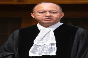 Ein Mann in einem schwarzen Gewand und weißem Hemd steht vor einer hölzernen Wand, trägt eine Brille und hat einen ernsten Gesichtsausdruck, was auf einen Richter hinweist.