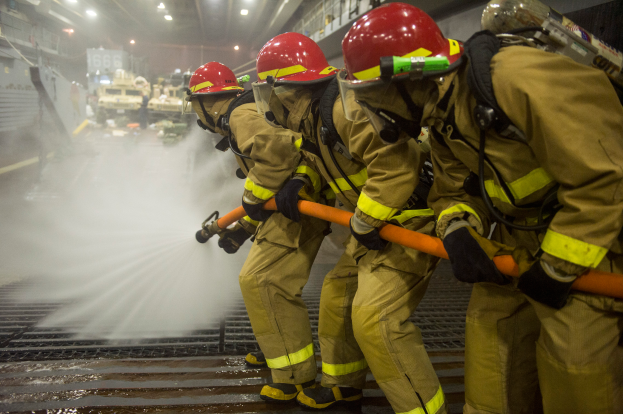 Feuerwehrleute in Helmen und Handschuhen spritzen Wasser aus Rohren auf ein Feuerwehrfahrzeug, mit verschiedenen Gegenständen und einem Boden im Hintergrund.