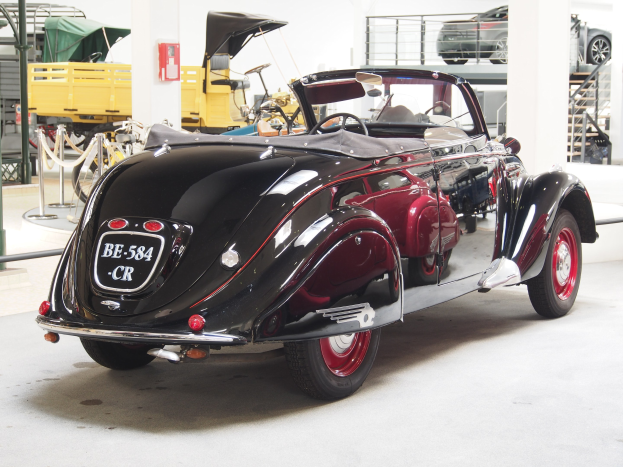 Schwarzes und rotes Auto in einer Garage mit anderen Fahrzeugen, Pfosten, Ketten und Gegenständen geparkt, mit einem Feuerlöscher an der Wand im Hintergrund.