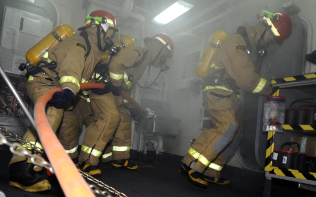 Feuerwehrleute in Schutzausrüstung mit Helmen und Sauerstoffflaschen arbeiten an einem Feuerwehrauto, mit einem Gestell voller Gegenstände und Hintergrundrohren und -lichtern.