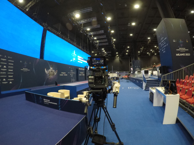 Eine Videokamera auf einem Stativ vor einem großen Bildschirm an einem Event mit Stühlen, Tischen, Bannern, Treppen, Geländern und Deckenleuchten.