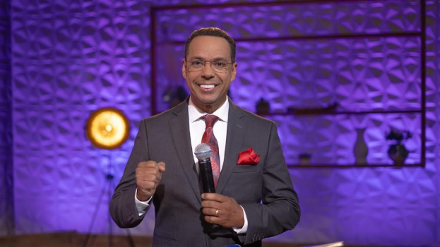 Stephen Colbert, Moderator von "The Daily Show mit Trevor Noah", steht auf einer Bühne und hält ein Mikrofon in der Hand, trägt einen Anzug, eine Krawatte und eine Brille, während im Hintergrund ein Scheinwerfer und ein Ständer zu sehen sind.