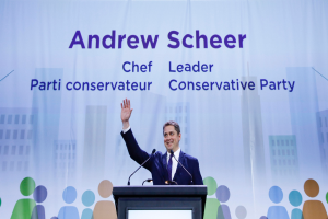 Andrew Scheer, der Vorsitzende der Konservativen Partei, steht hinter einem Podium mit zwei Mikrofonen und einem Bildschirm, mit einem Banner mit Text und Gebäudeillustrationen dahinter.