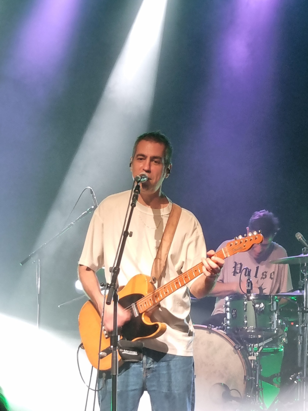 Ein Mann spielt eine E-Gitarre auf der Bühne, während er in ein Mikrofon singt, während ein anderer Musiker Schlagzeug spielt.