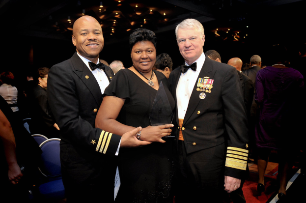 Zwei Männer in schwarzen Blazern und weißen Hemden stehen neben einer Frau in einem schwarzen Kleid, die eine Trophäe auf einem roten Teppich hält, während andere im Hintergrund sitzen und stehen unter beleuchteten Decken.