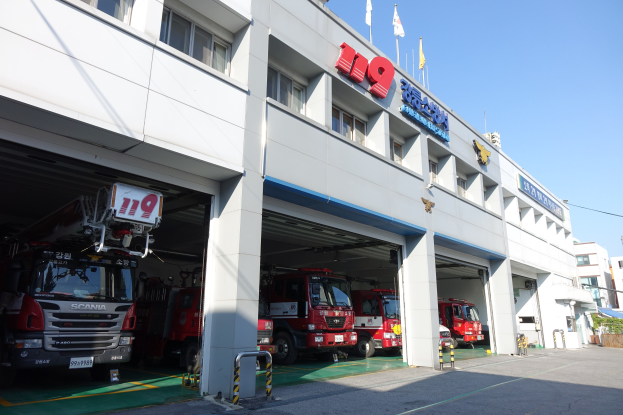 Feuerwache mit mehreren Feuerwehrautos vor der Fassade geparkt, mit Fenstern, Säulen, Namensschildern und Flaggen im Vordergrund; andere Gebäude und ein klarer blauer Himmel im Hintergrund.