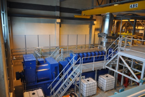 Fabrikinnensicht mit Trägern, einer Treppe mit Geländer, Containern, Deckenleuchten, freiliegenden Rohren und einer Wand.