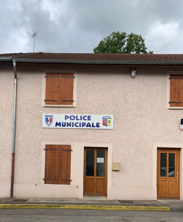 Ein Gebäude der Polizei municipale mit einem Schild, Fenstern, Türen, einer Namenskennzeichnung, einem Rohr, einem Baum und einem bewölkten Himmel im Hintergrund.