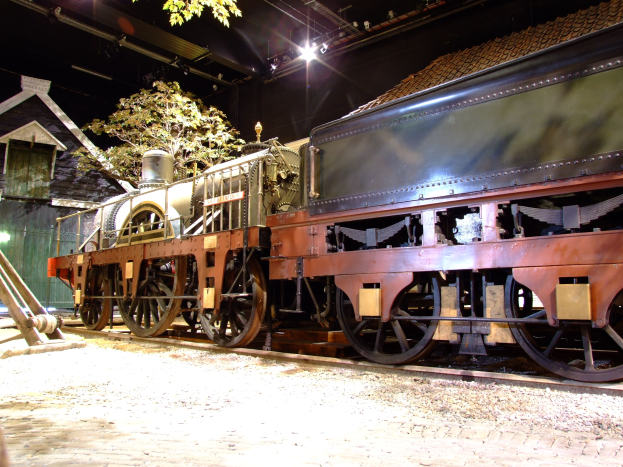 Eine schwarze Dampflokomotive in einem Museums Setting gezeigt, positioniert auf Schienen mit einem Backsteingebäude, Baum, Deckenbeleuchtung und Metallrahmen im Hintergrund.