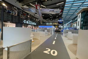 Ein geräumiger Raum mit weißen Wänden, einem aufgehängten Flugzeug, einem Geländer auf der linken Seite, verstreuten Gegenständen auf der rechten Seite, Texttafeln im Hintergrund und an Metallstangen befestigten Deckenleuchten, wahrscheinlich am Düsseldorfer Flughafen.