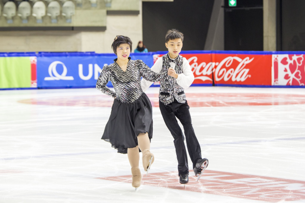 Zwei Eiskunstläufer, ein Mann und eine Frau, gleiten Hand in Hand über eine Eisfläche, mit Zuschauern und Event-Elementen im Hintergrund.