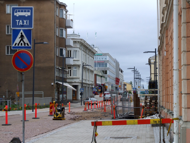 Eine Stadtstraße mit Gebäuden, Straßenlaternen, Verkehrsschildern, Absperrpollern, Autos, Baustelle mit Verkehrsschildern und einem Himmel mit Wolken.