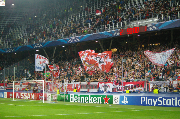 Fußballspiel in einem hell erleuchteten Stadion mit einer großen Menge, grünem Rasen, Torpfosten, Bannern und einem Bildschirm in der oberen linken Ecke.
