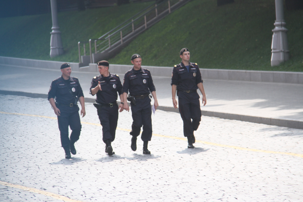 Drei Polizisten in Uniform und mit Mützen gehen eine Straße mit Pfosten, Geländern, Treppen und Gras im Hintergrund entlang.