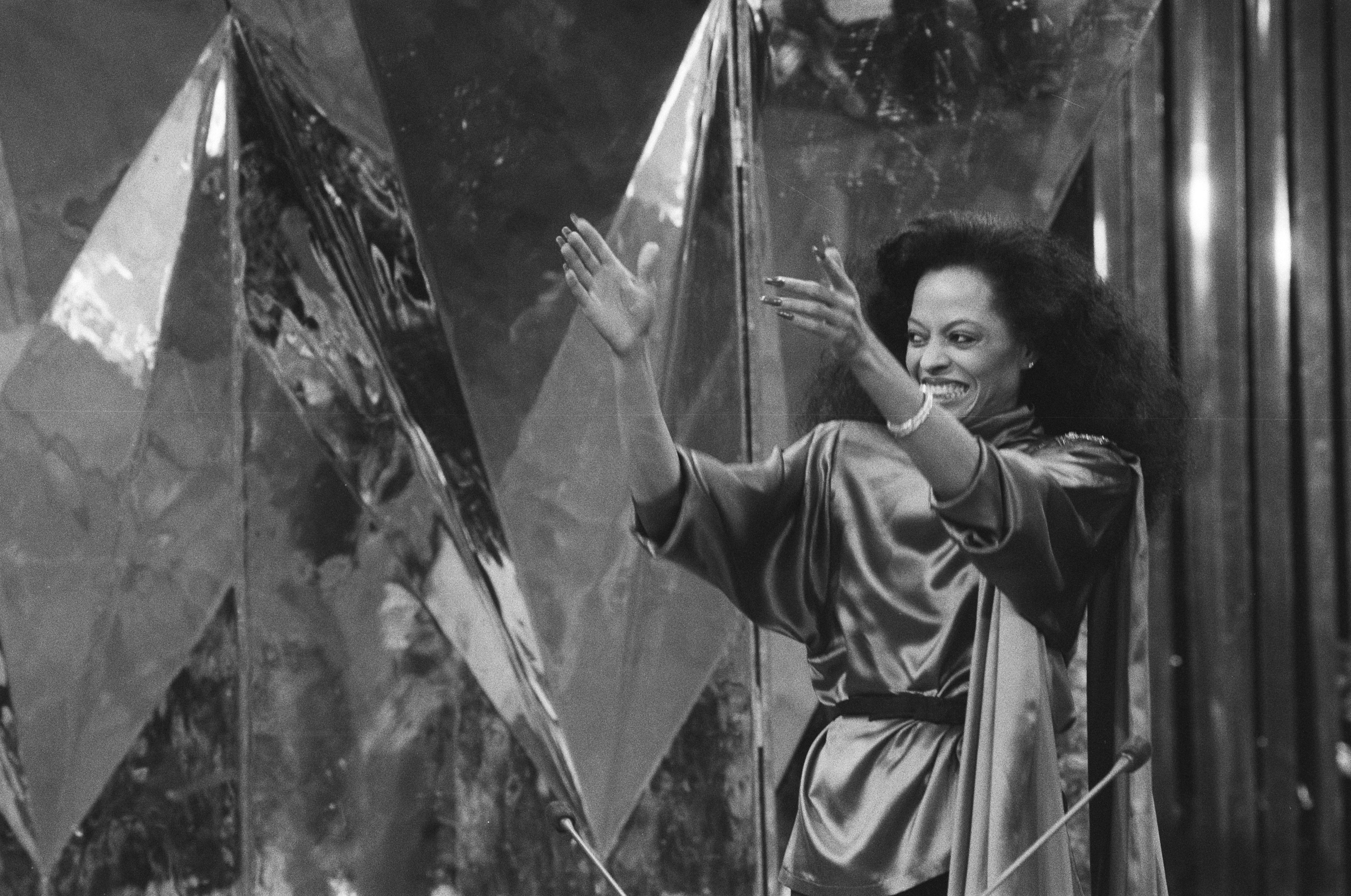 Schwarzes und weißes Foto von Diana Ross in einem weißen Kleid mit einem klassischen Bob, die ein Mikrofon hält und selbstbewusst vor einer Wand steht.