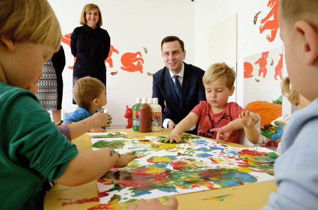 Kinder sitzen um einen Tisch mit Farbe an den Händen, beschäftigt mit einer kreativen Aktivität mit Papieren und Flaschen auf dem Tisch, während eine Frau im Hintergrund mit Gemälden an der Wand lächelt.