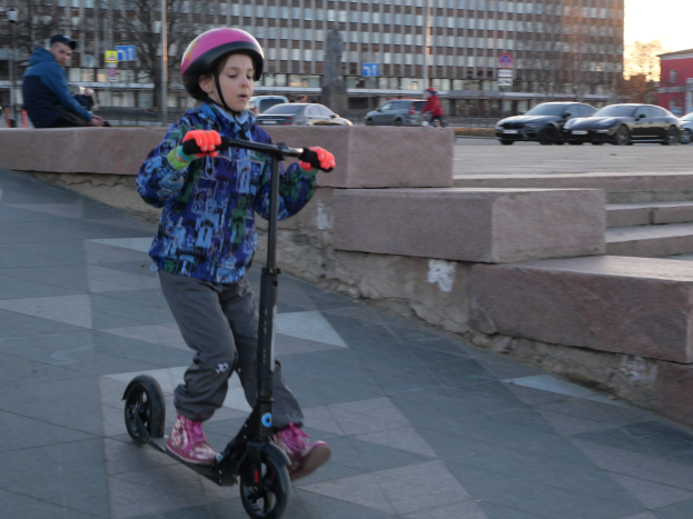Ein junger Junge in Helm und Handschuhen fährt einen Roller eine Treppe hinunter, mit Fahrzeugen, Menschen, Bäumen, Pfählen, Brettern, Gebäuden und einem klaren blauen Himmel im Hintergrund.