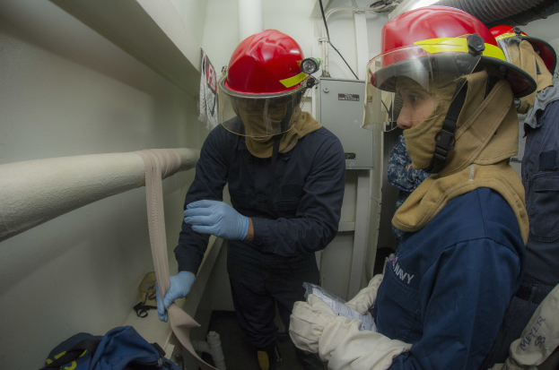 Drei Personen in Feuerbekämpfungsausrüstung stehen zusammen in einem Raum mit Rohren, einer Tasche, Maschinen und Beleuchtung im Hintergrund.