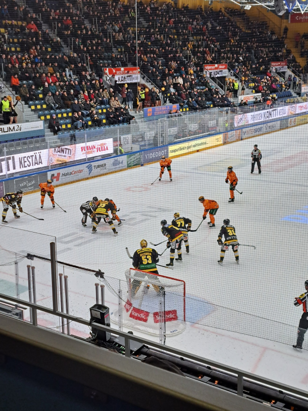 Eine Gruppe von Menschen, die Hockey auf einem Eisring spielt, mit einem Netz und einem umgebenden Zaun, Zuschauern im Hintergrund und Bannern mit Text, unter Stadionbeleuchtung.