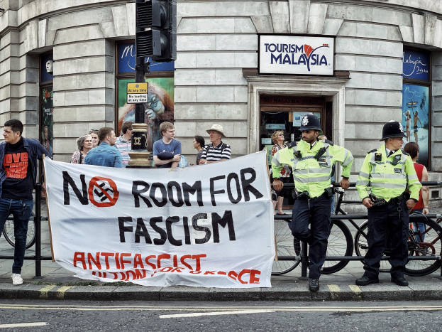 Eine Gruppe von Menschen mit einer 'Kein Platz für Faschismus'-Plakette steht vor zwei Polizisten auf einem Gehweg, mit Fahrrädern, einer Ampel und einem Gebäude im Hintergrund.