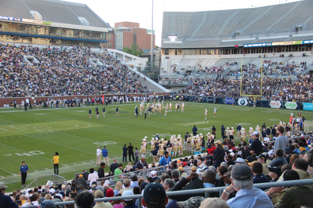 Ein Fußballspiel im Gange mit Spielern auf dem Feld und Zuschauern in den Rängen unter einem klaren blauen Himmel.