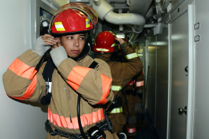 Feuerwehrleute in Uniform, die während einer Übung in einem Raum stehen, mit Rohren und Ausrüstung im Hintergrund.
