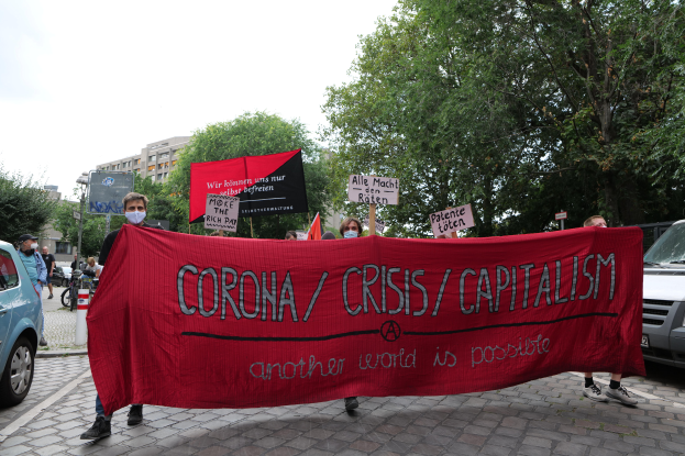 Eine Gruppe von Menschen marschiert mit einer roten Fahne, auf der "Corona-Krise/Kapitalismus" steht, eine Straße entlang, mit geparkten Fahrzeugen auf beiden Seiten und Gebäuden, Bäumen und einem klaren blauen Himmel im Hintergrund.