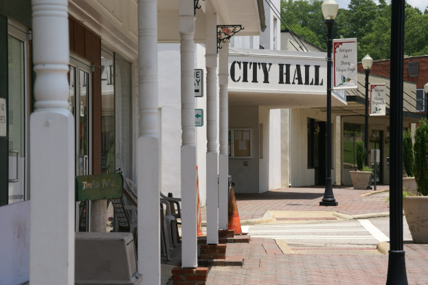 Ein Rathaus mit Gebäuden, Straßenmasten, Straßenlampen, Namenschildern, Kraftfahrzeugen, Absperrgittern, Stühlen, Hauspflanzen, Bäumen und einem bewölktem Himmel im Hintergrund.