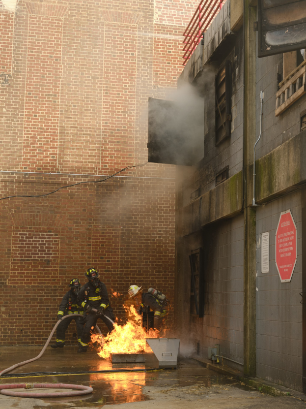Feuerwehrleute mit Helmen verwenden Schläuchen, um ein Gebäude Feuer zu löschen, während Rauch aufsteigt; benachbarte Gebäude mit Fenstern und ein Schild sind sichtbar.