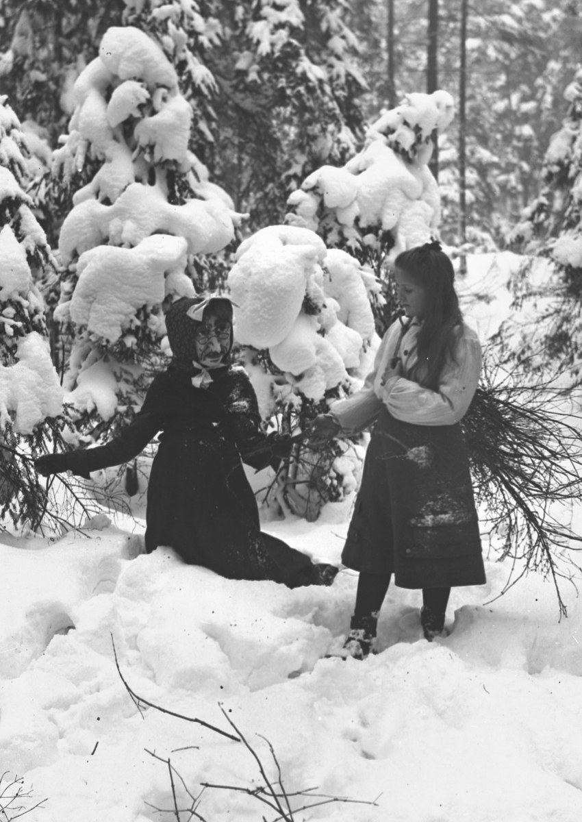 Zwei Kinder spielen in einem verschneiten Waldgebiet, umgeben von schneebedeckten Bäumen, in Schwarz-Weiß.