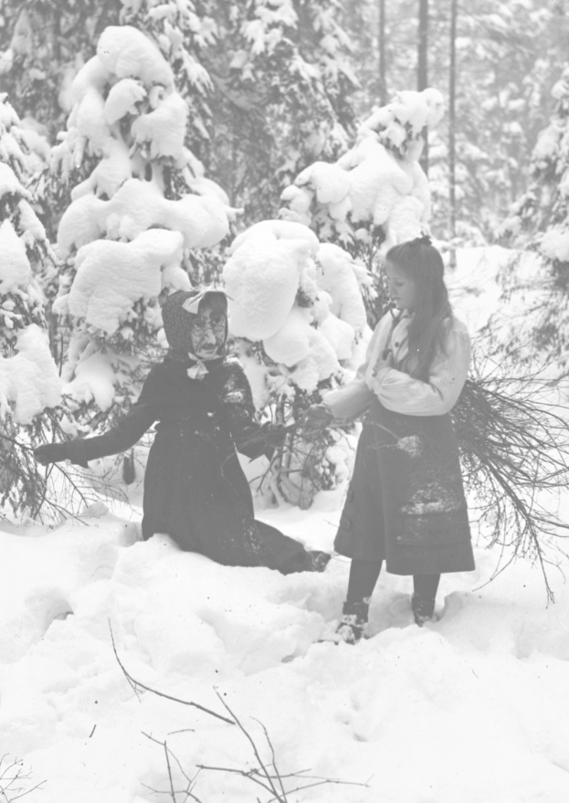 Zwei Kinder spielen in einem verschneiten Waldgebiet, umgeben von schneebedeckten Bäumen, in Schwarz-Weiß.