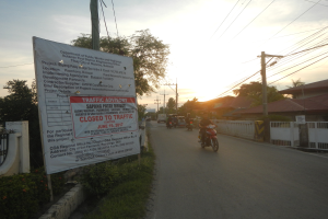 Gruppe von Menschen auf Motorrädern auf einer Straße mit einem "Verkehrshinweis Gesperrt für den Verkehr"-Schild, gesäumt von einer blumenbedeckten Wand, mit Häusern, Bäumen und einem klaren Himmel im Hintergrund.