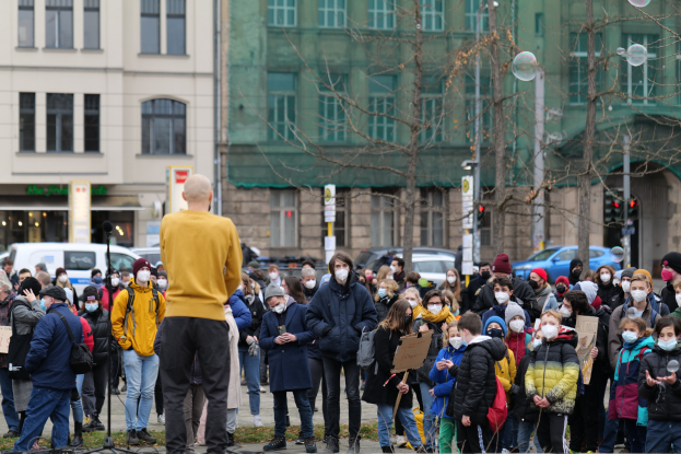 Eine Gruppe von Menschen demonstriert in Berlin, Deutschland, wobei einige Schilder halten und Masken tragen, vor einer Menge, mit einem Mikrofonständer im Vordergrund und Gebäuden, Bäumen und Fahrzeugen im Hintergrund.