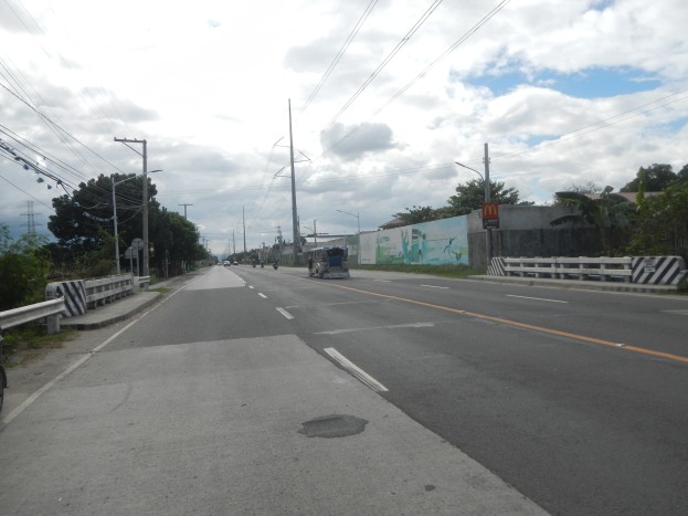 Arterielle Straße mit fahrenden Fahrzeugen, einem geparkten Motorrad, Geländern, Strommasten mit Kabeln, Bäumen, Gebäuden und einem bewölkten Himmel im Hintergrund.