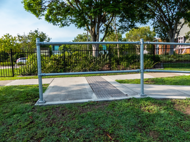 Metallzaun in einer grasbewachsenen Area mit umliegenden Pflanzen und Bäumen, ein Auto auf der Straße, Gebäude im Hintergrund und einen klaren blauen Himmel.